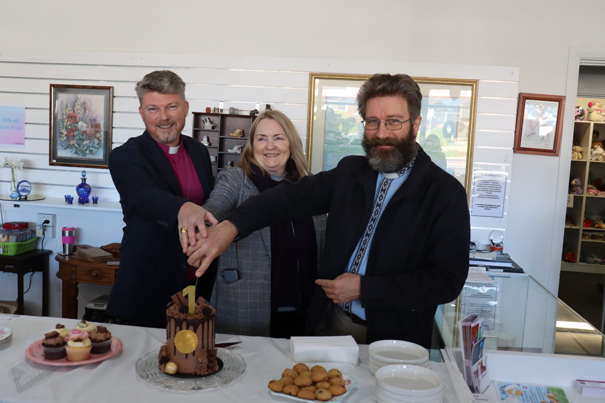 Bishop of Gippsland Dr Richard Treloar, volunteer coordinator Lee-Anne Croucher and Rector John Webster cut the 1 year birthday cake for the Drouin Anglican Church opportunity shop